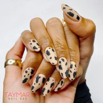 Uñas ovaladas beige con diseño de vaca en negro y un anillo dorado.