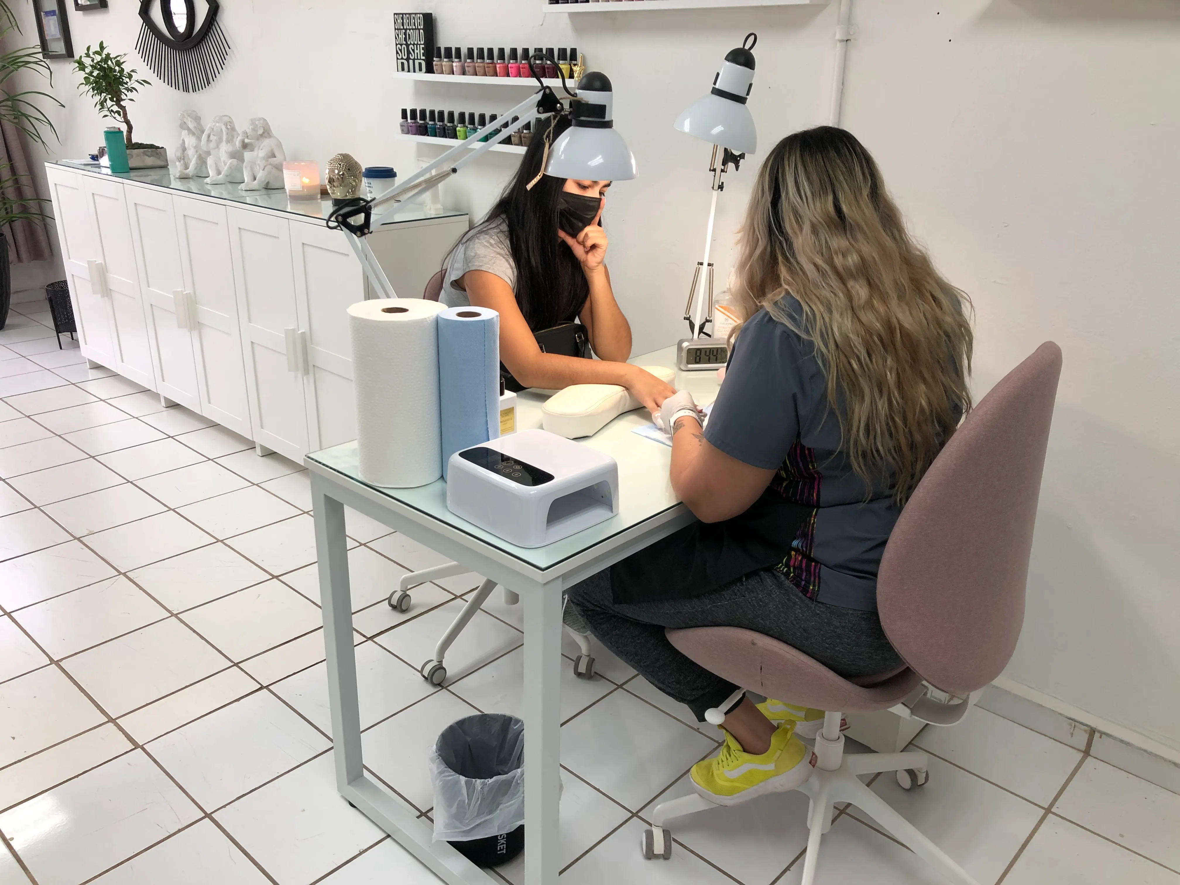 Una técnica de uñas haciendo una manicura a una clienta en una mesa blanca, con lámparas y esmaltes al fondo.