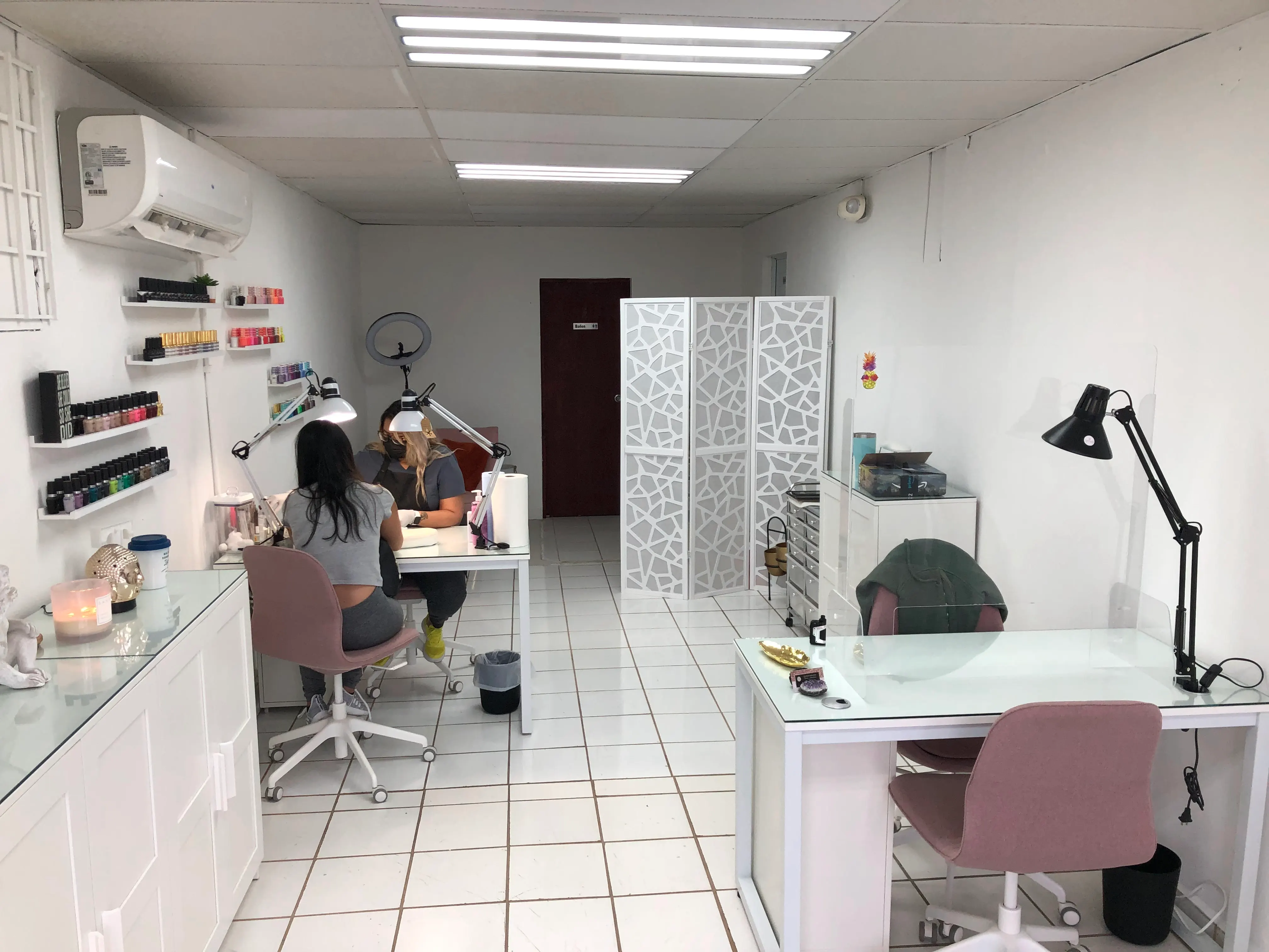 Interior del estudio: dos mesas de manicura, estantes de esmaltes y piso blanco.